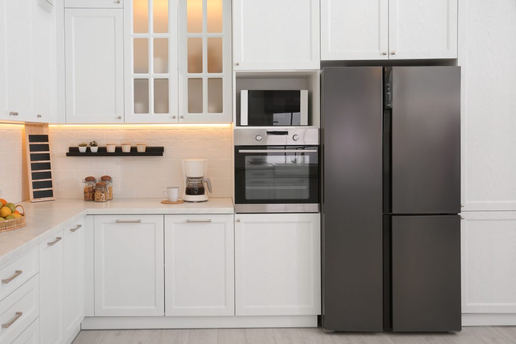 A tall four-door refrigerator standing at the end of a white shaker kitchen, its doors closed, a built-in oven and microwave in the run beside it
