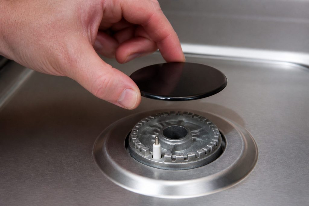 A hand lifting the cap of a gas burner straight up off its head, the igniter and ports exposed beneath