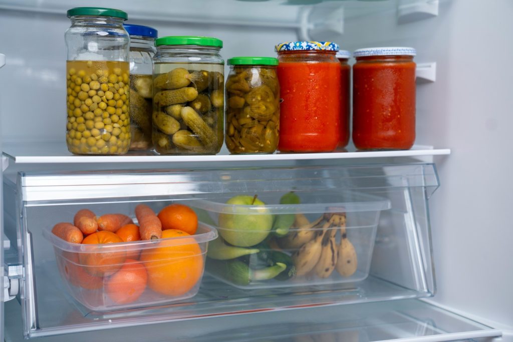 Preserving jars on a glass shelf above a crisper drawer of fruit and vegetables