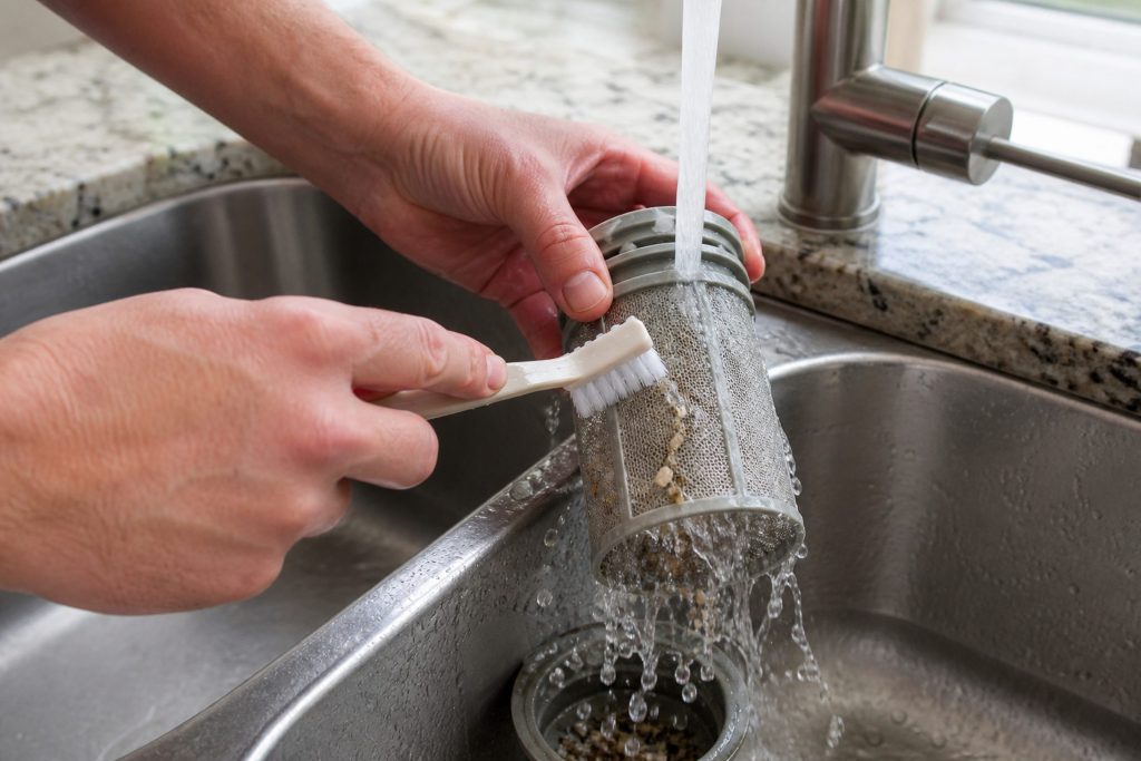 A dishwasher filter cylinder held under a running kitchen tap and rinsed in both hands