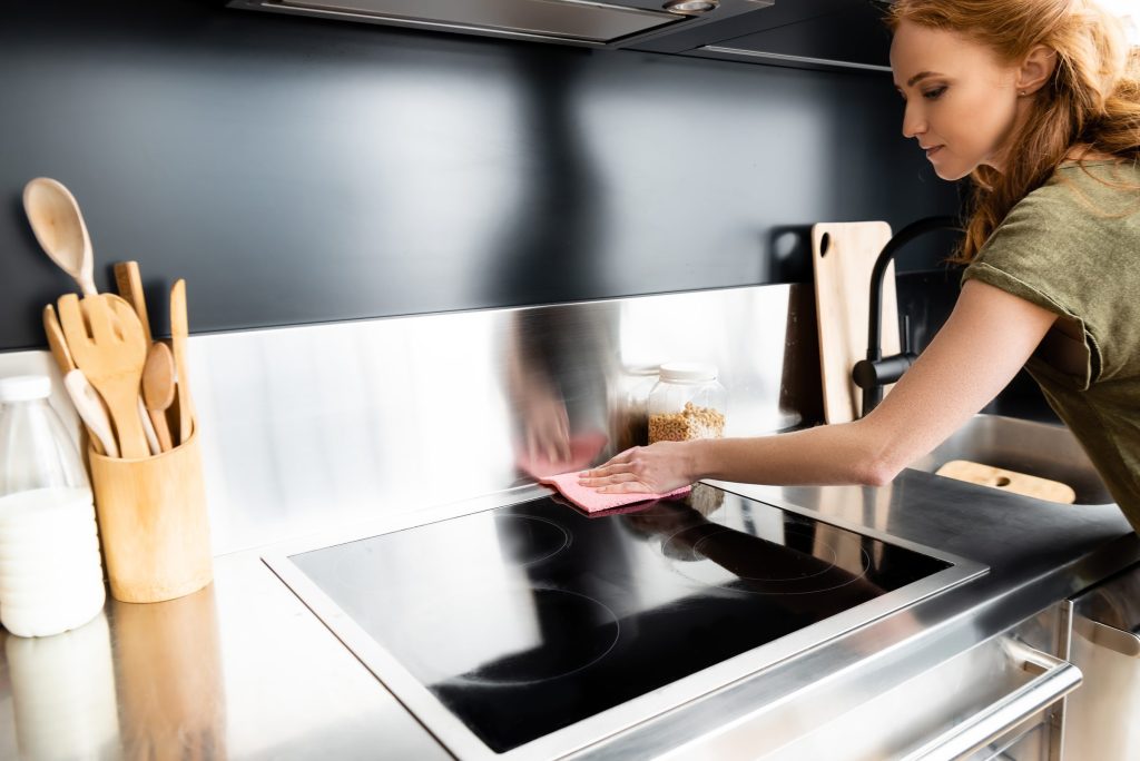 A woman wiping a black glass hob with a pink cloth in a dark kitchen.