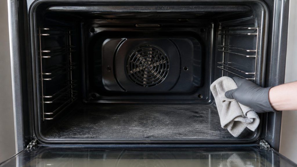 A gloved hand wiping out the enamel cavity of an oven with a cloth, the fan cover at the back