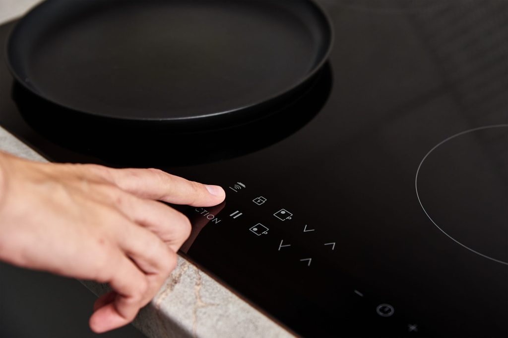 A finger on the touch control strip of an induction cooktop, a pan on the zone behind