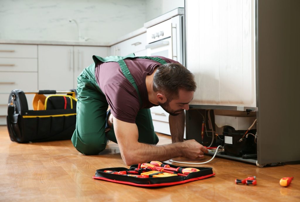 A technician kneeling at the base of a refrigerator with the lower panel off and tools laid out