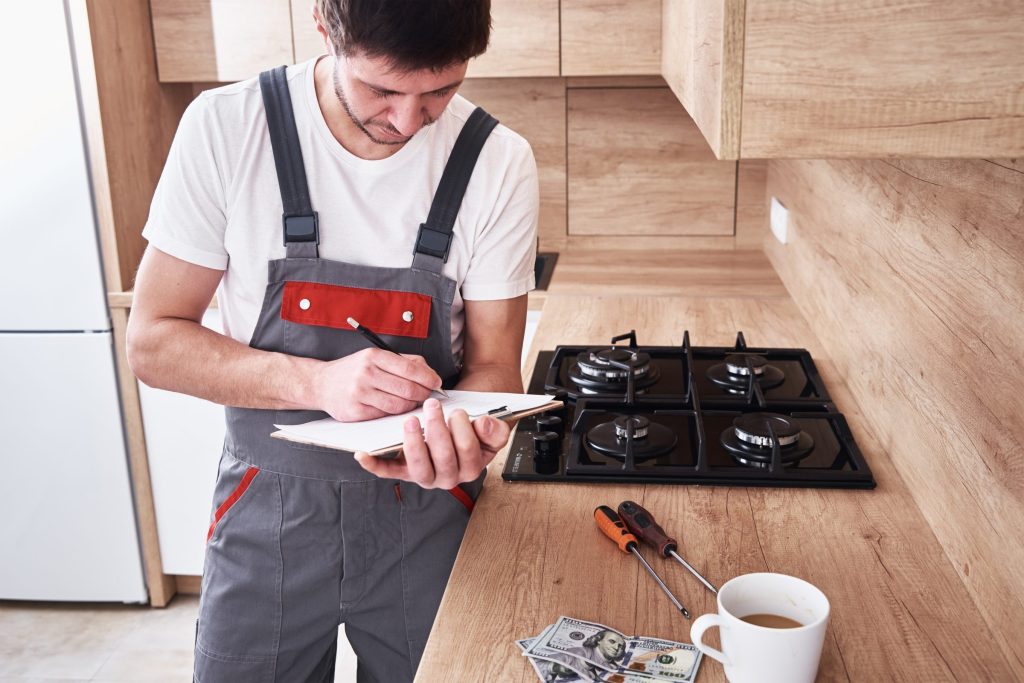 A technician writing on a pad beside a gas cooktop, tools and a cup on the worktop