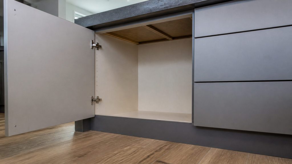A cabinet door standing open beneath a dark stone kitchen island, showing the empty space and the underside of the counter above, a stack of drawers pushed close on one side.