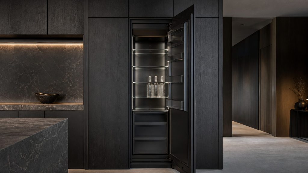 A tall full-height refrigerator column standing open in a dark luxury kitchen, showing glass shelves and a very dark interior lining lit softly from within, a few plain glass bottles on one shelf.