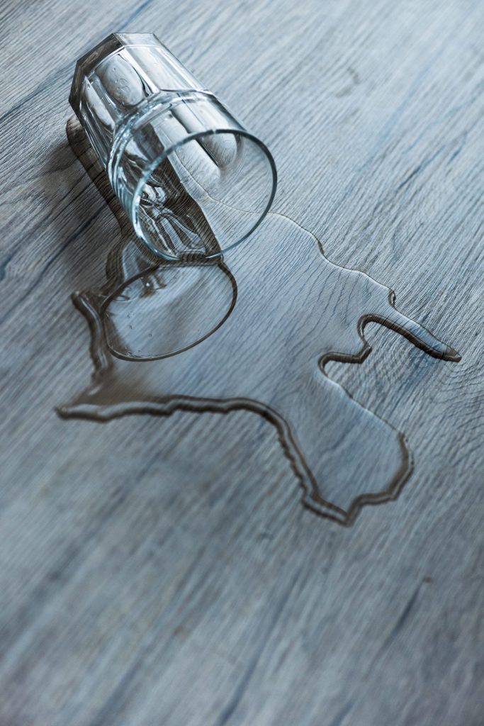 A tipped drinking glass with water spreading across a pale wood floor