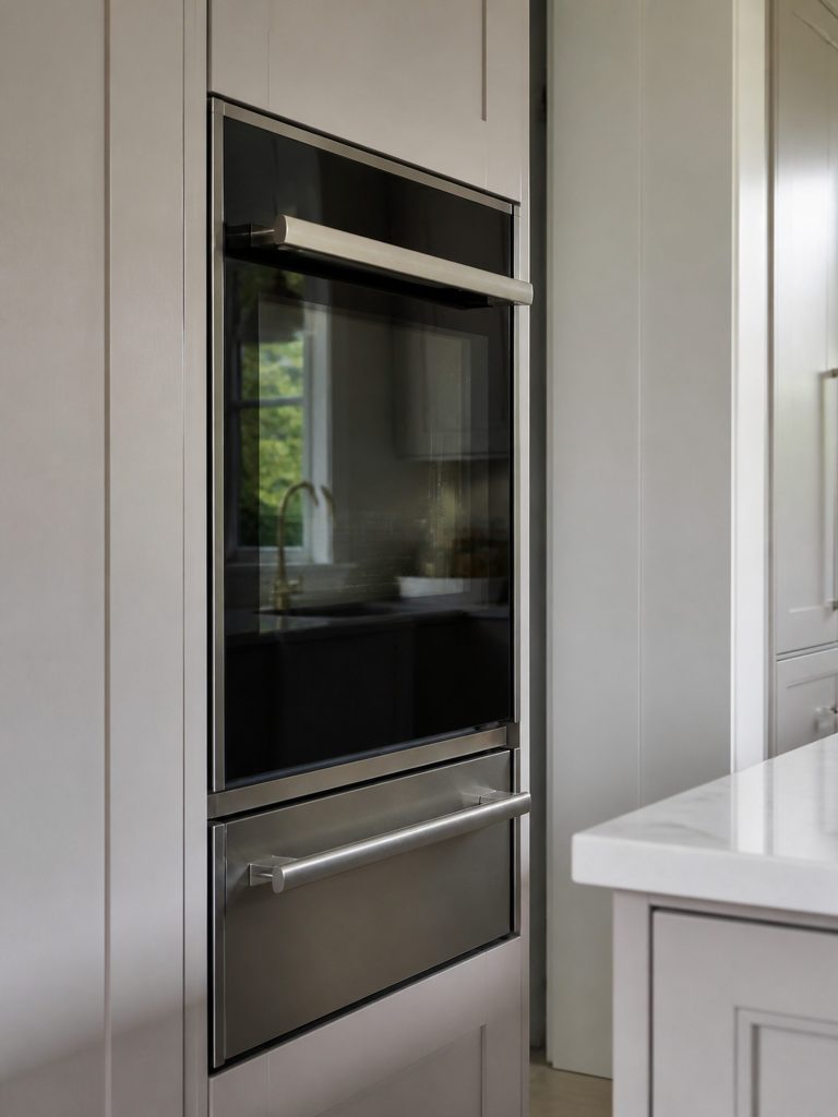 A narrow built-in oven in a tall cabinet column above a warming drawer, door closed, dark glass