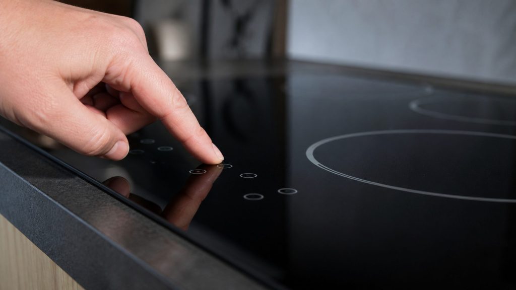 The touch control strip of an electric cooktop photographed close and at a low angle, a finger about to rest on it, the elements dark behind.