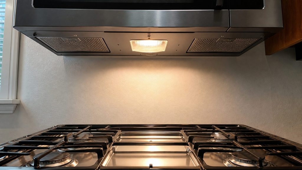 The underside of a microwave hood combination with its cooktop light on, casting a bright pool over the burners of the range below.