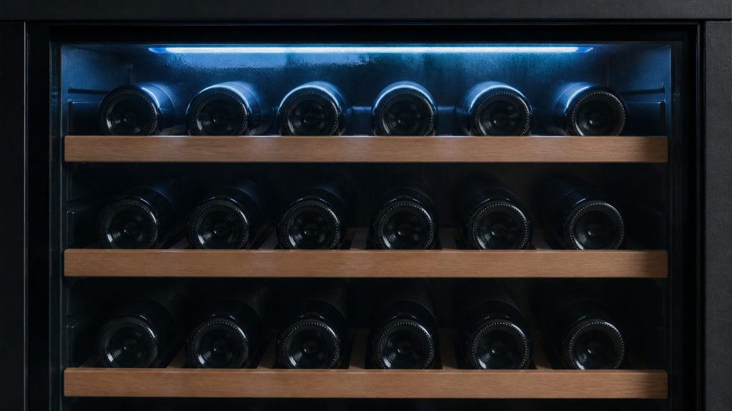 Rows of dark bottles on wooden runners behind glass, a cool light strip along the top