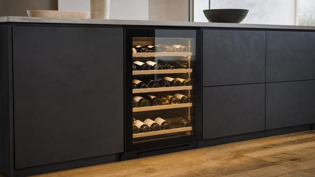 An undercounter wine cabinet built into dark cabinetry, bottles lying on wooden shelves behind a glass door, lit warm inside