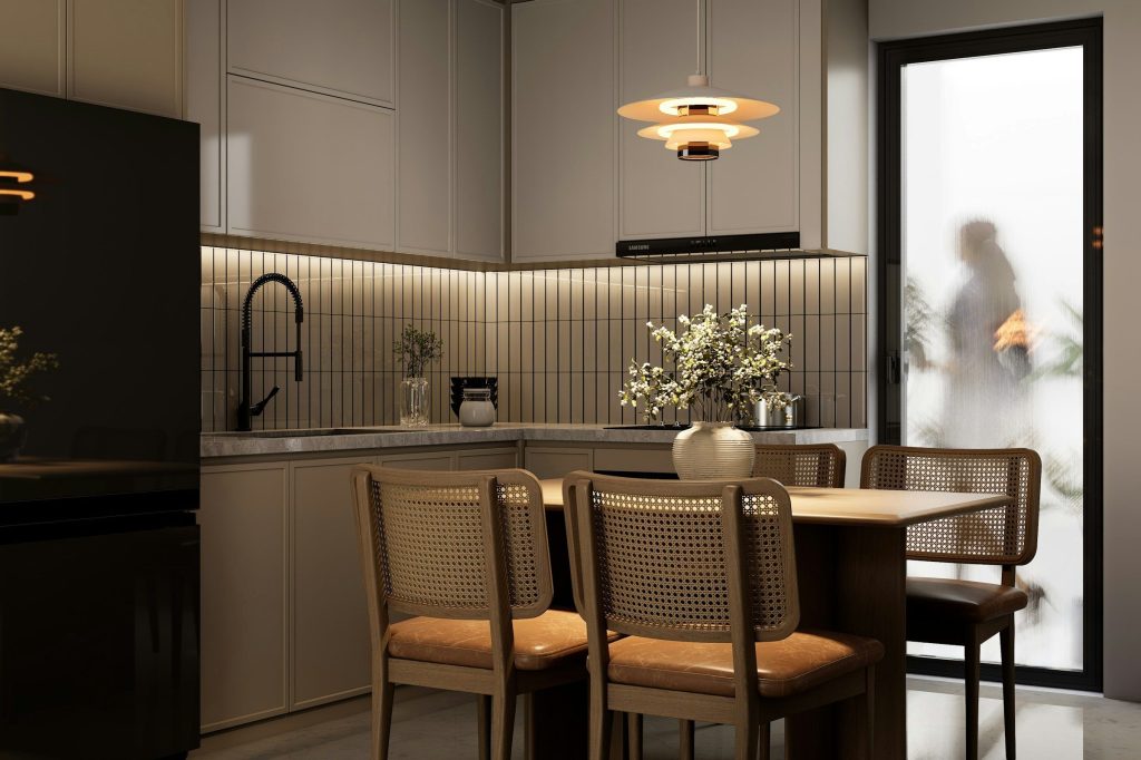A pale kitchen seen past a dining table with cane-backed chairs, a pendant light above it and daylight coming through a glazed door