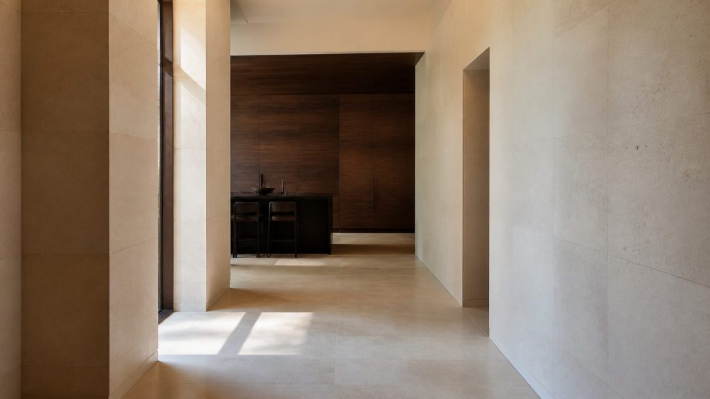 A wide hallway of pale stone opening into a dark wood kitchen at the far end, warm daylight from one side, tall cabinetry visible beyond.