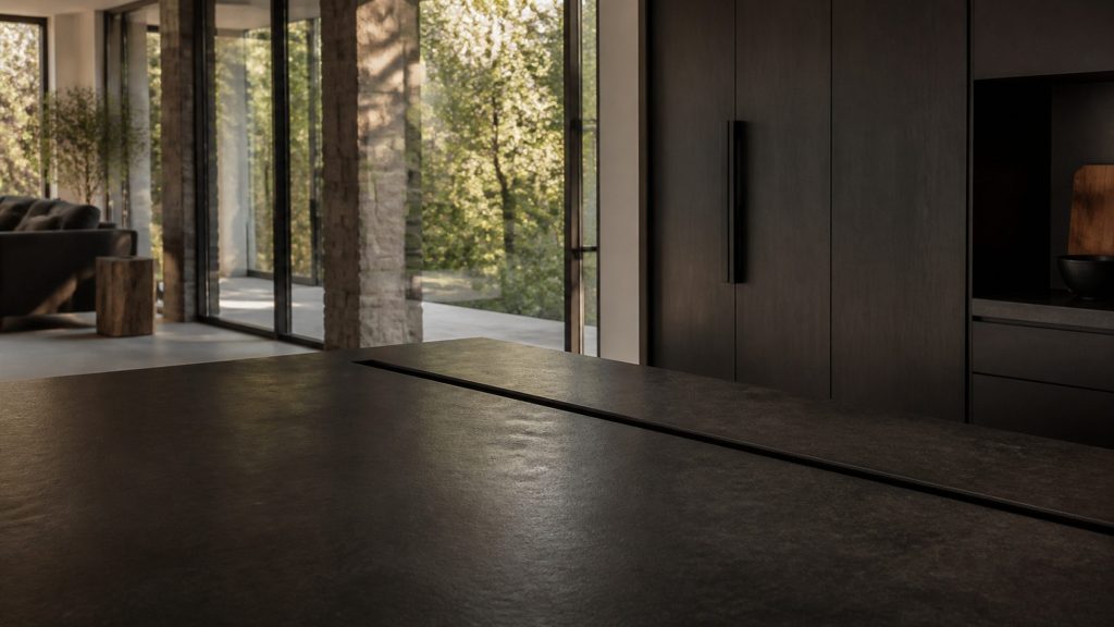 A cooking island in a dark open-plan kitchen seen from a few paces back, a low stone-topped counter with a slot running along its far edge, warm daylight from tall windows and greenery outside.