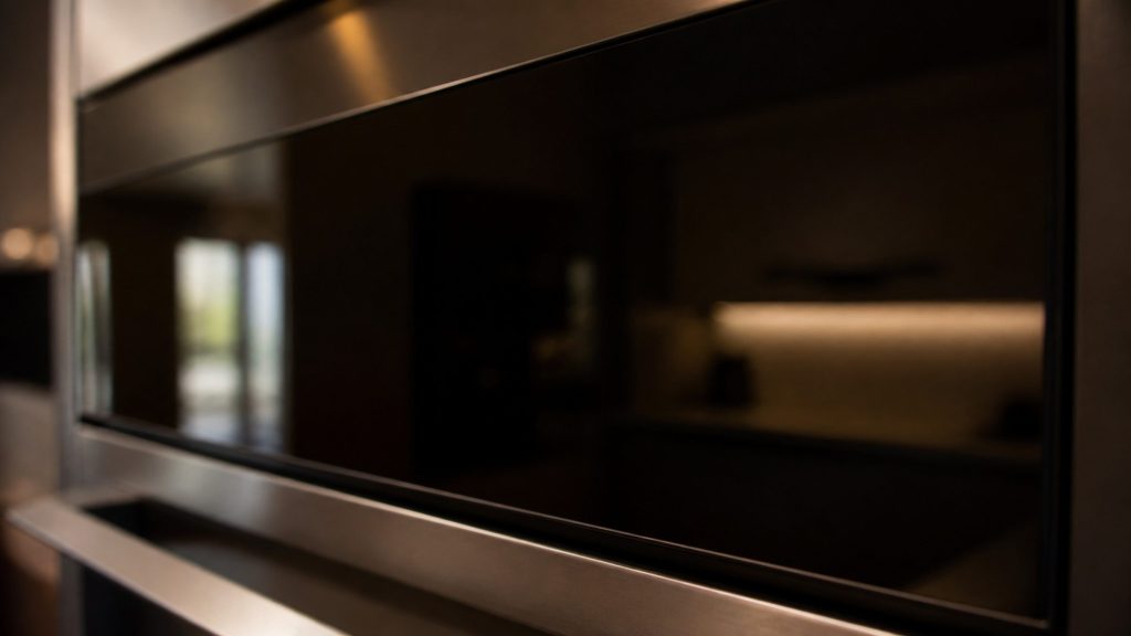 A very close, slightly angled view of a dark glass control fascia in a kitchen, its surface reflecting warm room light, everything beyond it thrown out of focus.