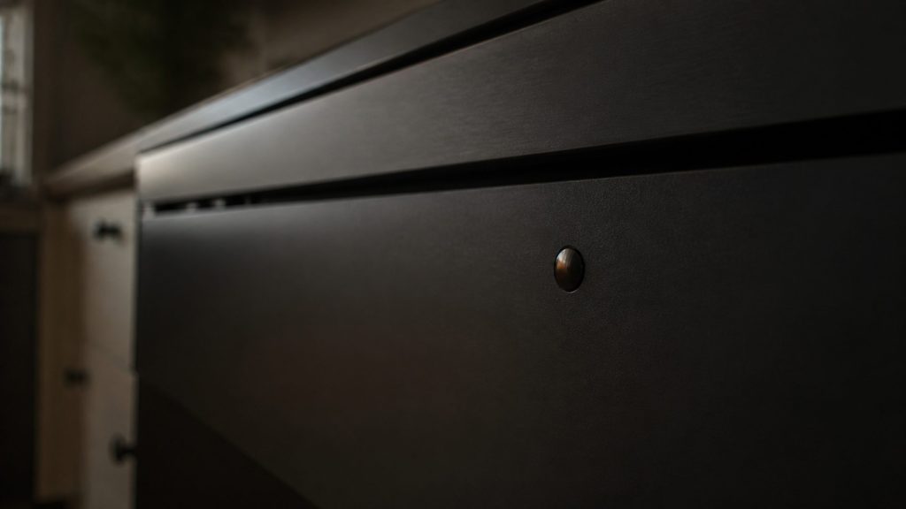 A close view along a dark appliance fascia in a shadowed kitchen, one small unlit indicator lens catching a warm reflection, the rest of the surface plain.