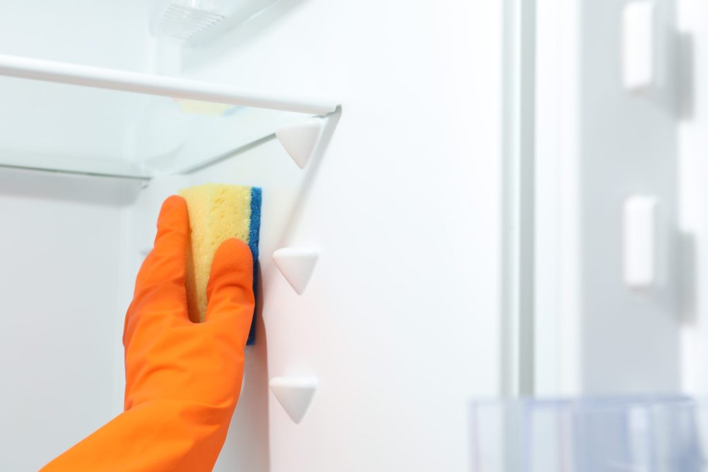 A gloved hand wiping down the interior shelf and door fittings of a refrigerator