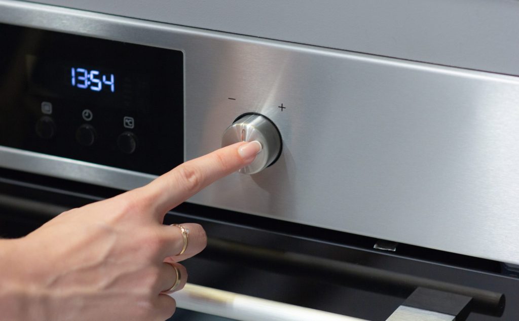 A finger pressing the control dial beside the lit display of a built-in oven