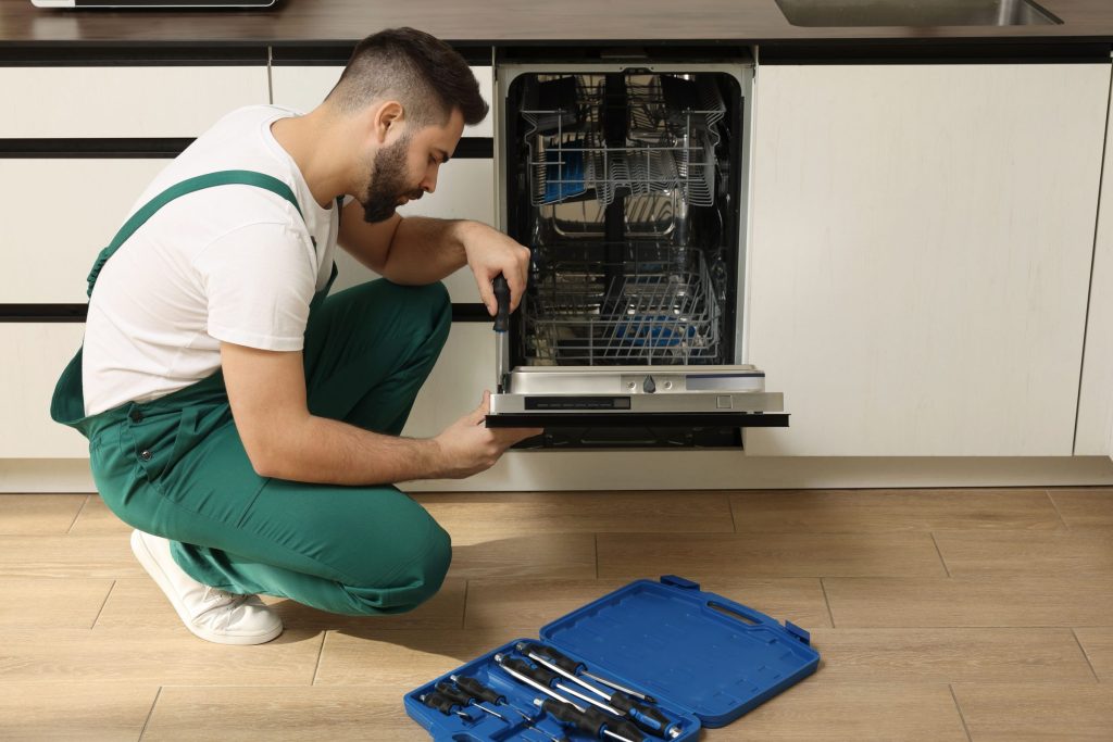 A technician crouched at the lowered door of a built-in dishwasher, tool roll open on the floor