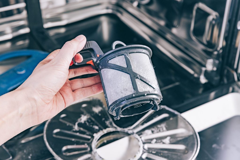 A hand lifting the cylindrical filter out of the floor of a dishwasher tub