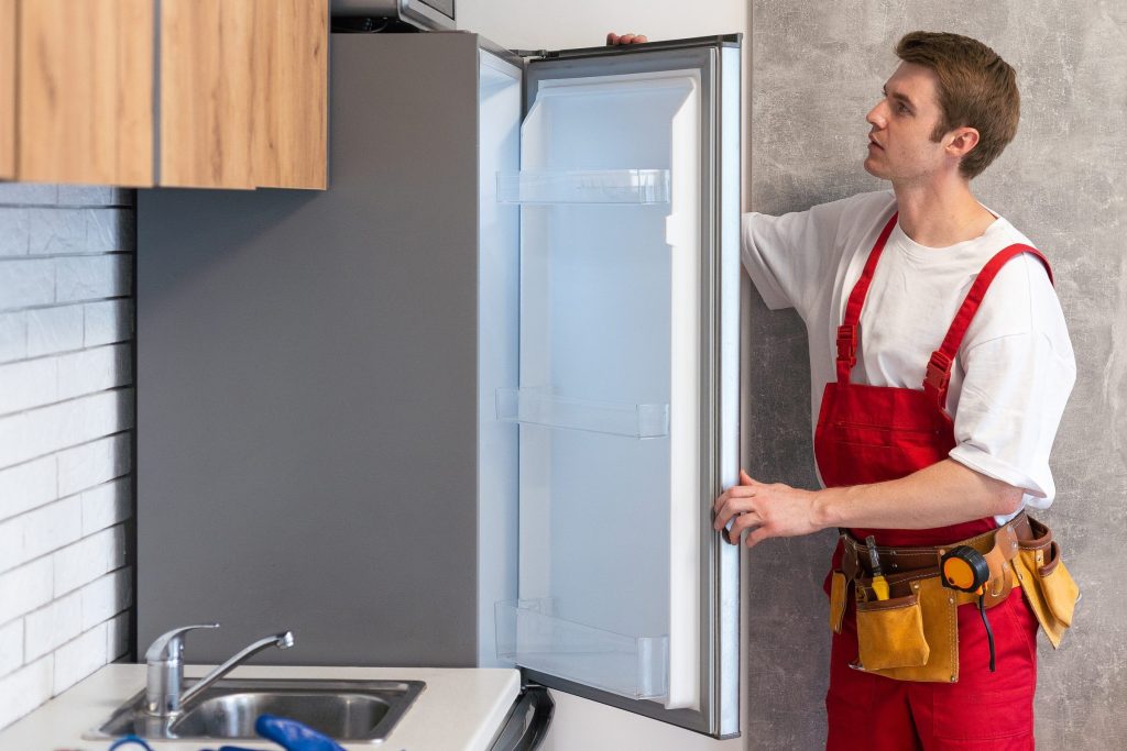 A technician holding open the door of a tall refrigerator beside a kitchen sink