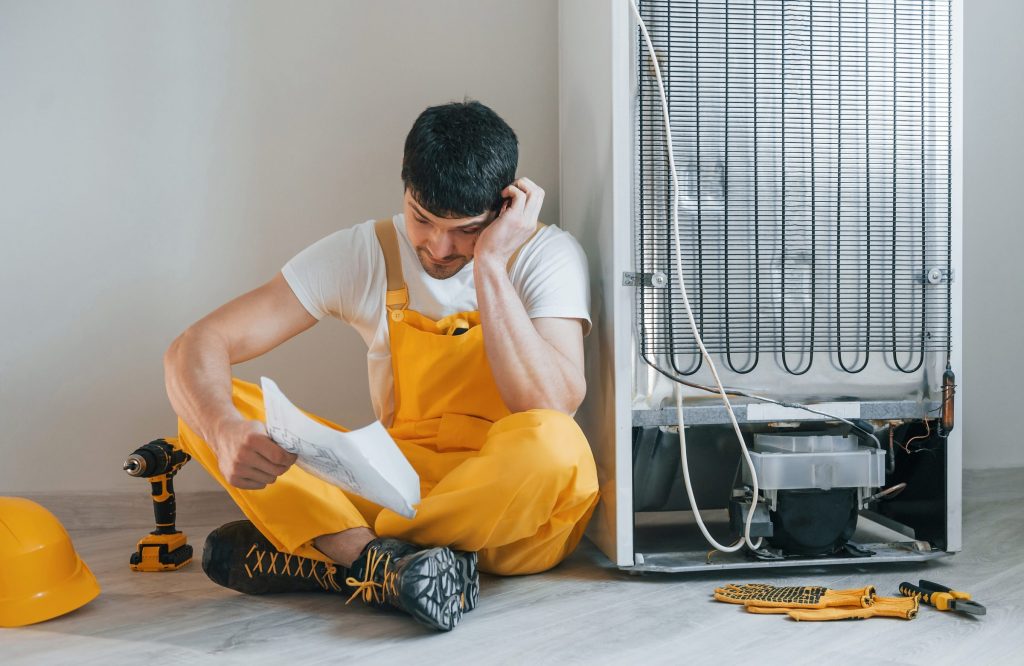A technician sitting at the back of a refrigerator with the rear panel off and the compressor exposed