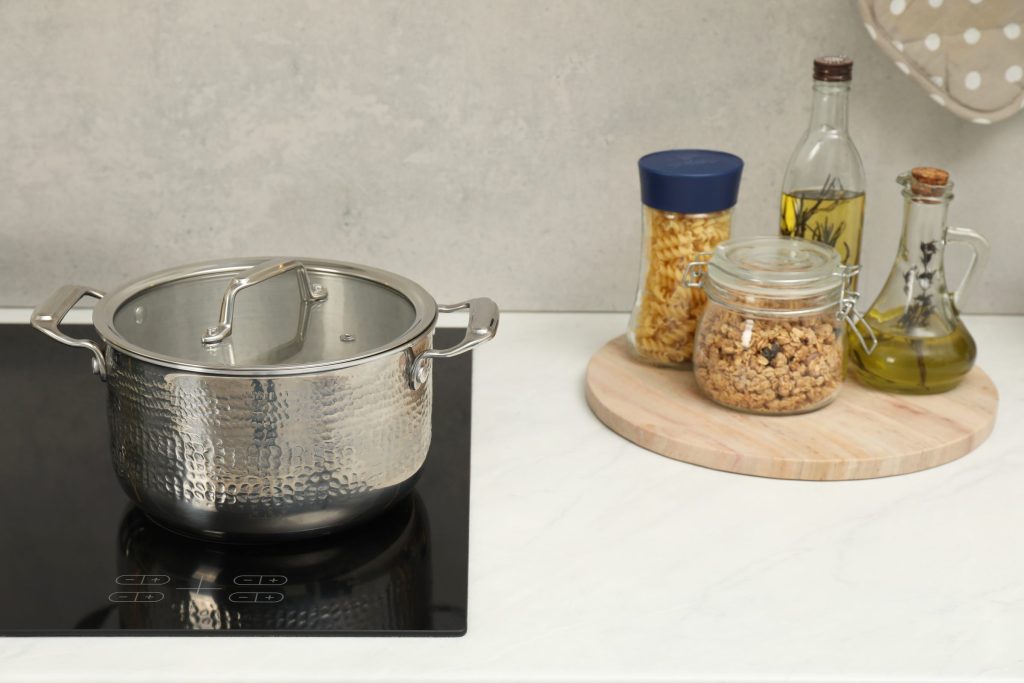A stainless pot on a glass cooktop with jars and a board on the counter behind