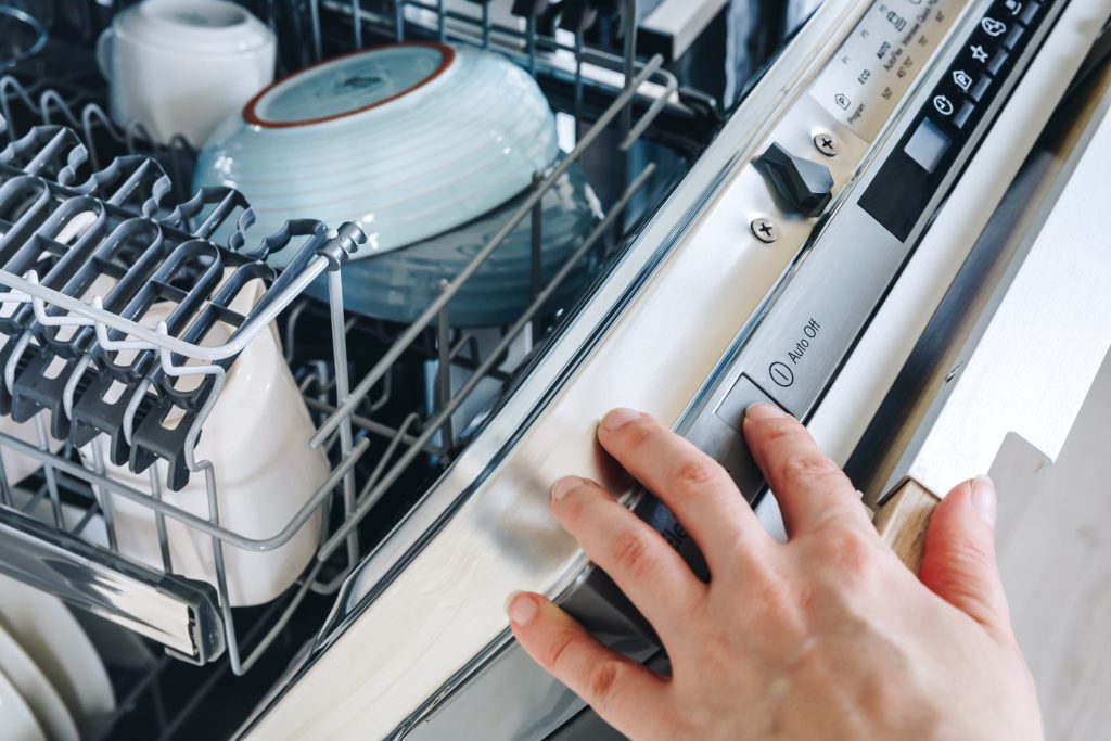 A hand at the control panel on the top edge of a dishwasher door